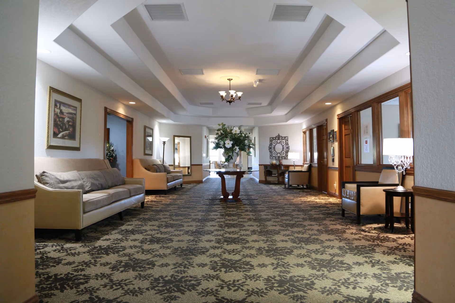 A spacious, well-lit hallway in a senior living facility with patterned carpet, beige sofas and armchairs along the walls, a central wooden table with a large floral arrangement, framed artwork on the walls, and ceiling lights.