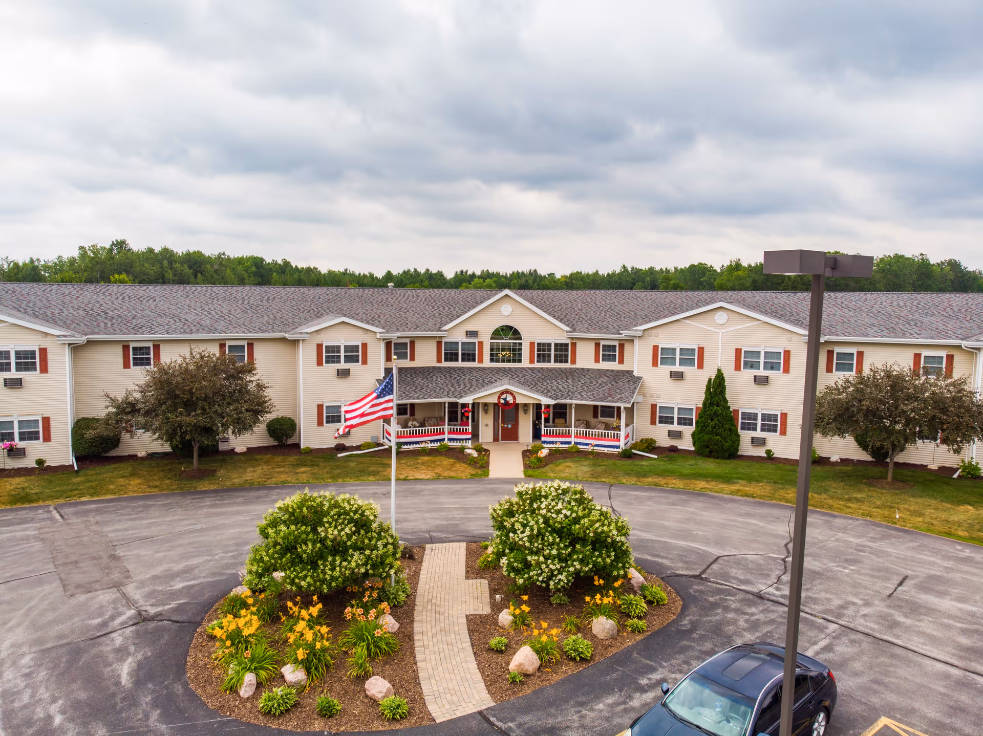 Front exterior of a two-story senior living building with a circular driveway, landscaped island, and an American flag in front.