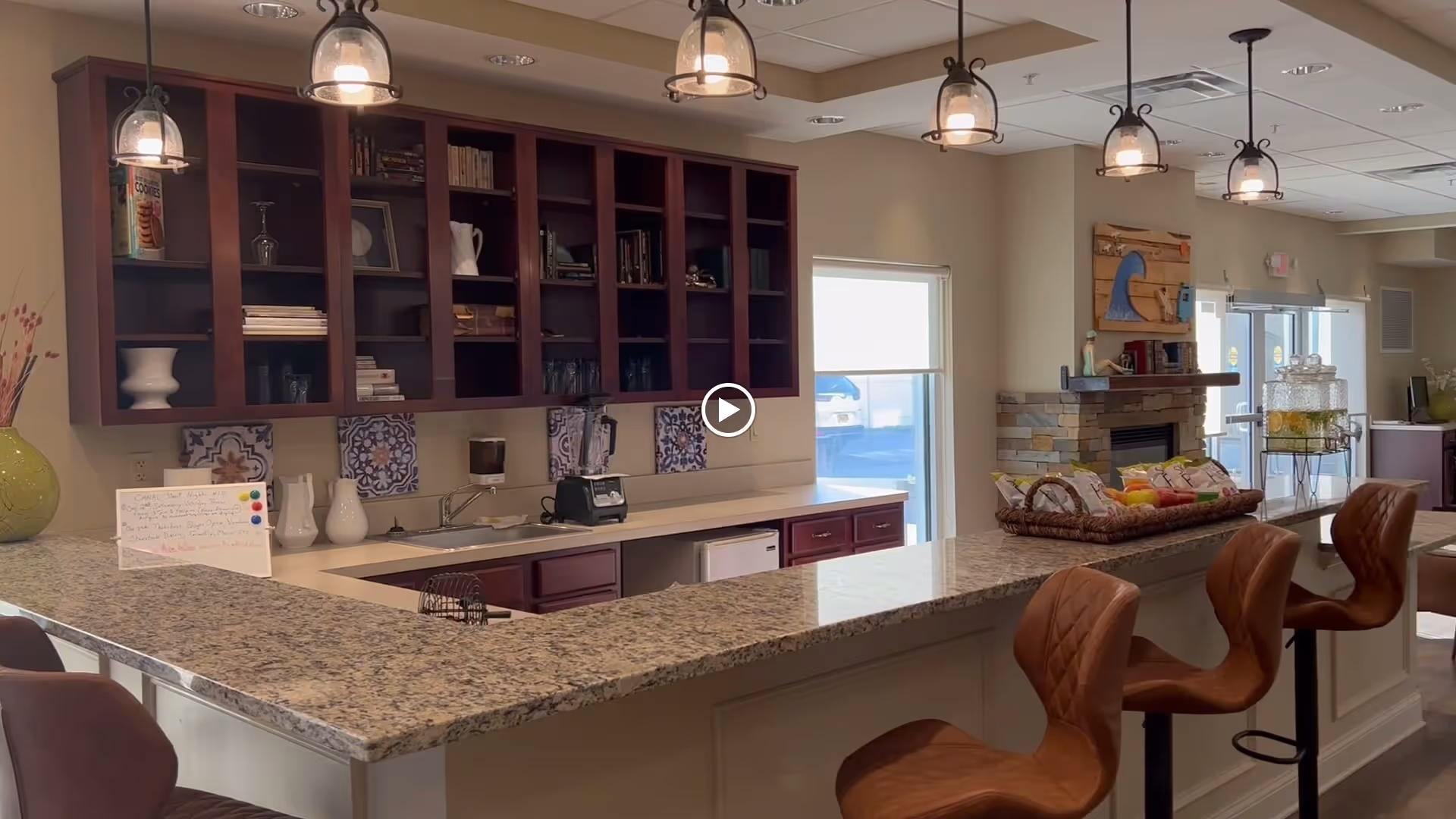 Interior view of a senior living facility's kitchen and common area featuring a granite countertop bar with three brown leather bar stools. Above the counter are hanging pendant lights. Behind the counter are dark wood cabinets with open shelves holding books and decorative items. A blender and other kitchen items are on the countertop. In the background, there is a stone fireplace with a wooden mantel and a large window letting in natural light.