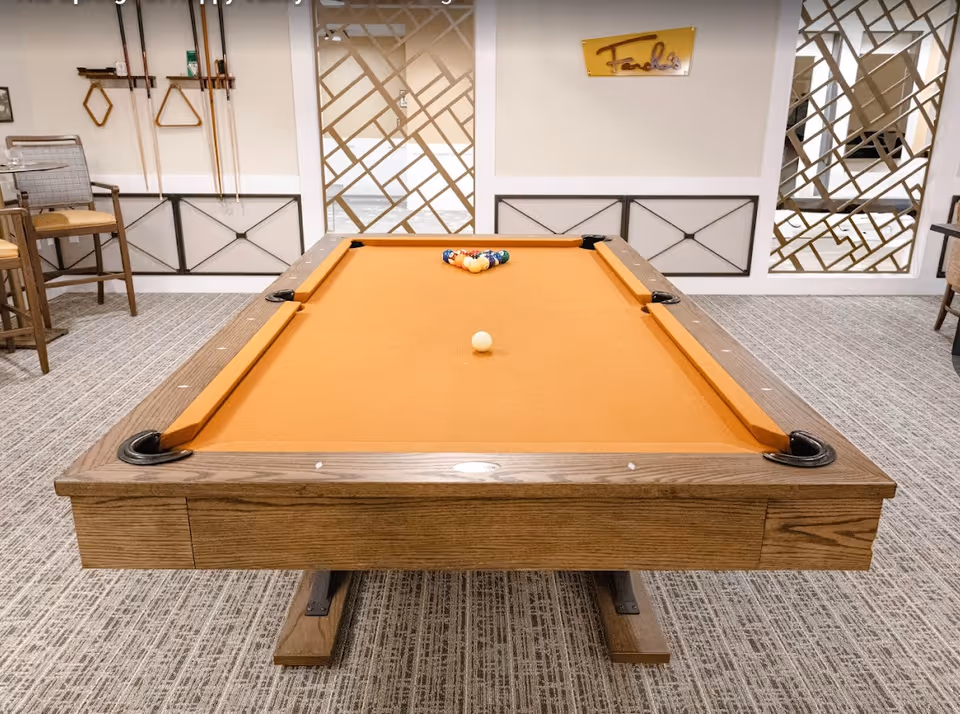 A billiards table with orange felt set up for play in a carpeted recreation room with cue racks and seating.