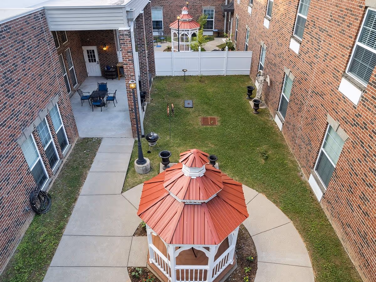 Outdoor courtyard area at Kempton of Hermitage featuring a red-roofed white gazebo in the center, surrounded by a concrete walkway, green grass, brick building walls with multiple windows, a patio area with a table and chairs, a barbecue grill, and decorative statues near the building.