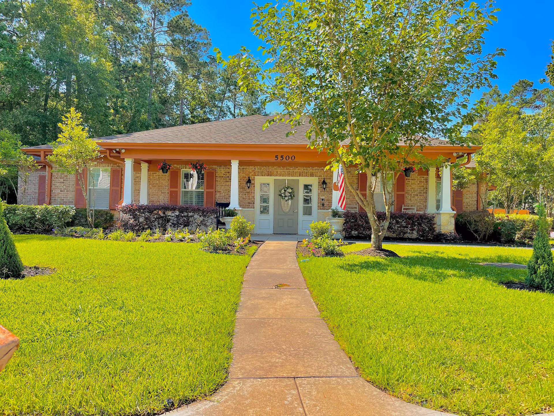 Front view of a single-story brick cottage with a covered porch, central walkway, manicured lawn and trees.