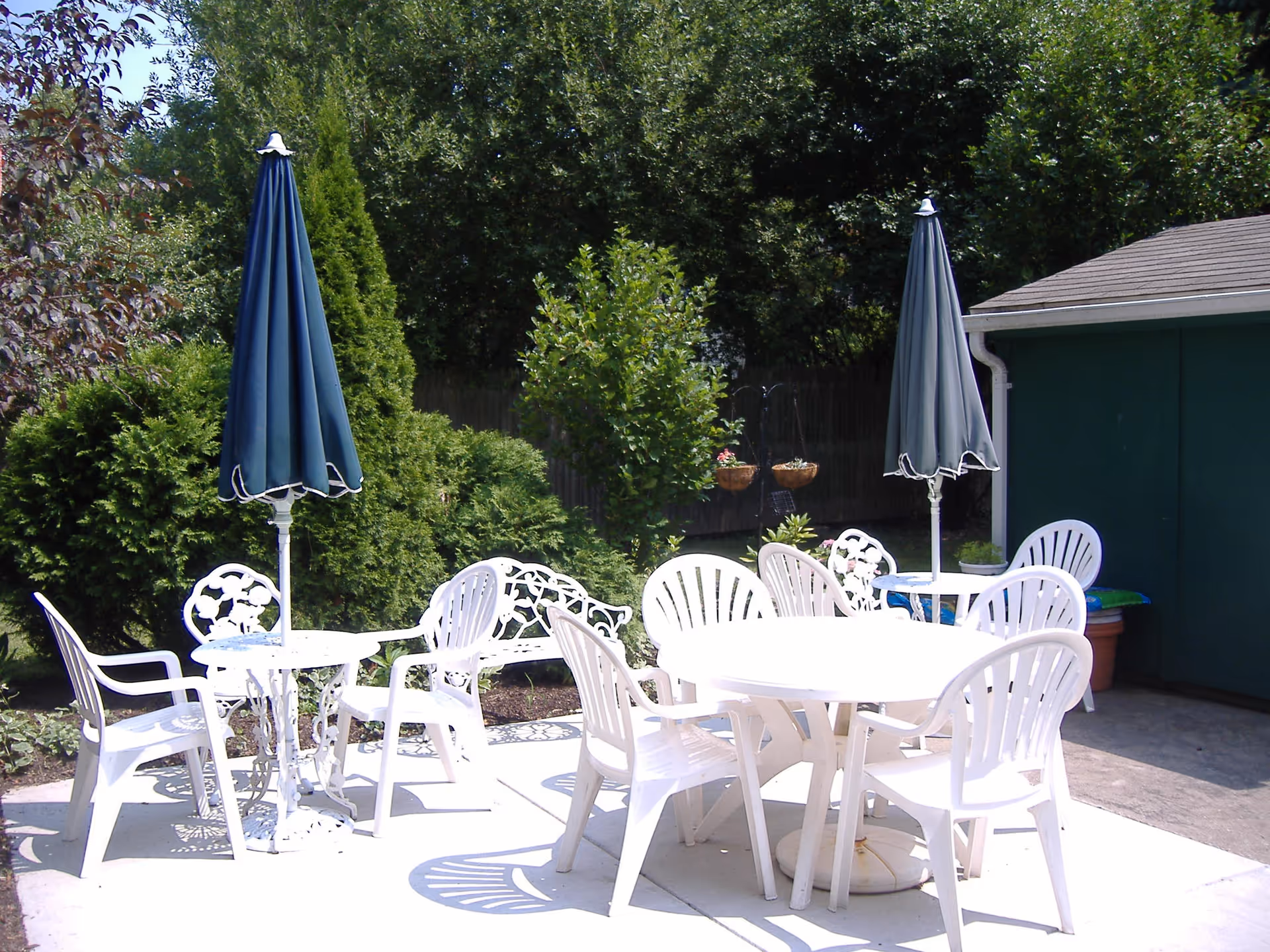 Outdoor patio area with white plastic chairs and tables, two closed blue umbrellas, surrounded by green bushes and trees, next to a green building.