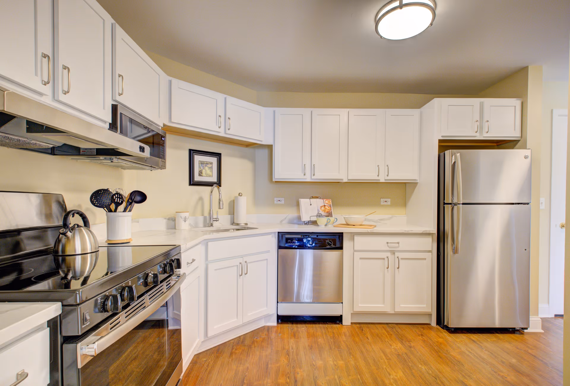 Modern kitchen with white cabinets, stainless steel appliances including a refrigerator, dishwasher, and stove with a kettle on it. The kitchen has wooden flooring and a ceiling light fixture.