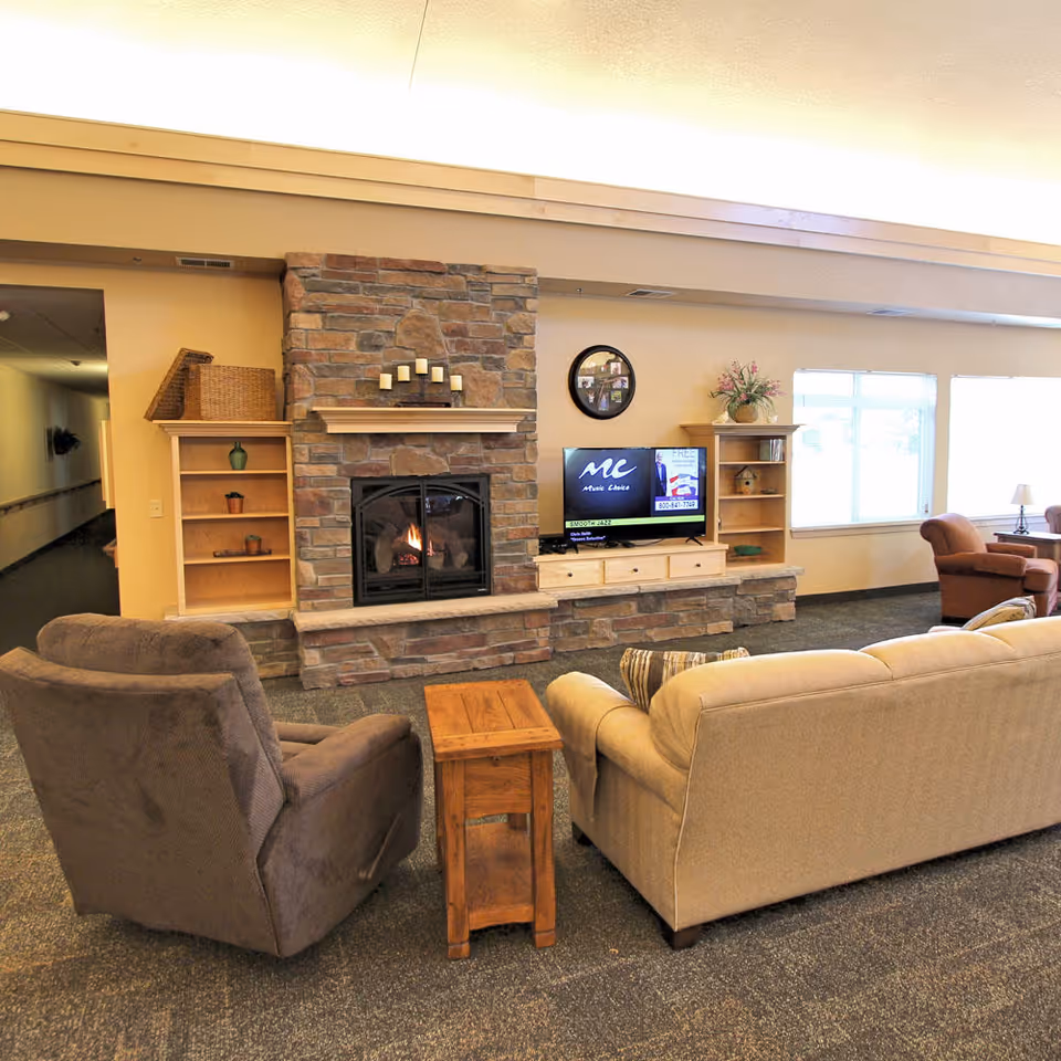 A cozy living room area in an assisted living facility featuring a stone fireplace with lit candles on the mantle, a flat-screen TV on a wooden cabinet, a beige sofa, a brown armchair, a small wooden side table, and large windows letting in natural light.