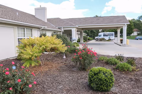 Exterior view of Vintage Park at Hiawatha showing a landscaped garden with bushes and flowers in front of a beige building with a covered entrance. A white shuttle bus is parked near the entrance under the covered area.
