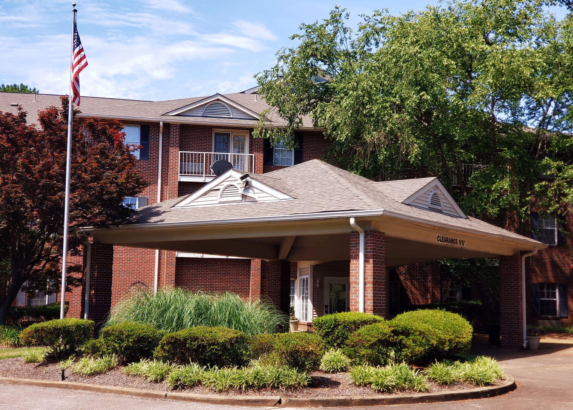 Exterior view of a senior living facility named Kennington Pointe featuring a brick building with multiple windows, a covered entrance with clearance sign, well-maintained bushes and trees, and an American flag on a flagpole.