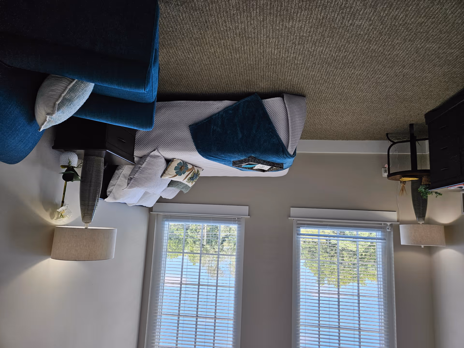 A cozy living room with a blue armchair, a white couch with decorative pillows, a dark wooden side table with a lamp and a flower vase, two large windows with white blinds letting in natural light, a dark wooden TV stand with a flat-screen TV, and a beige carpeted floor.