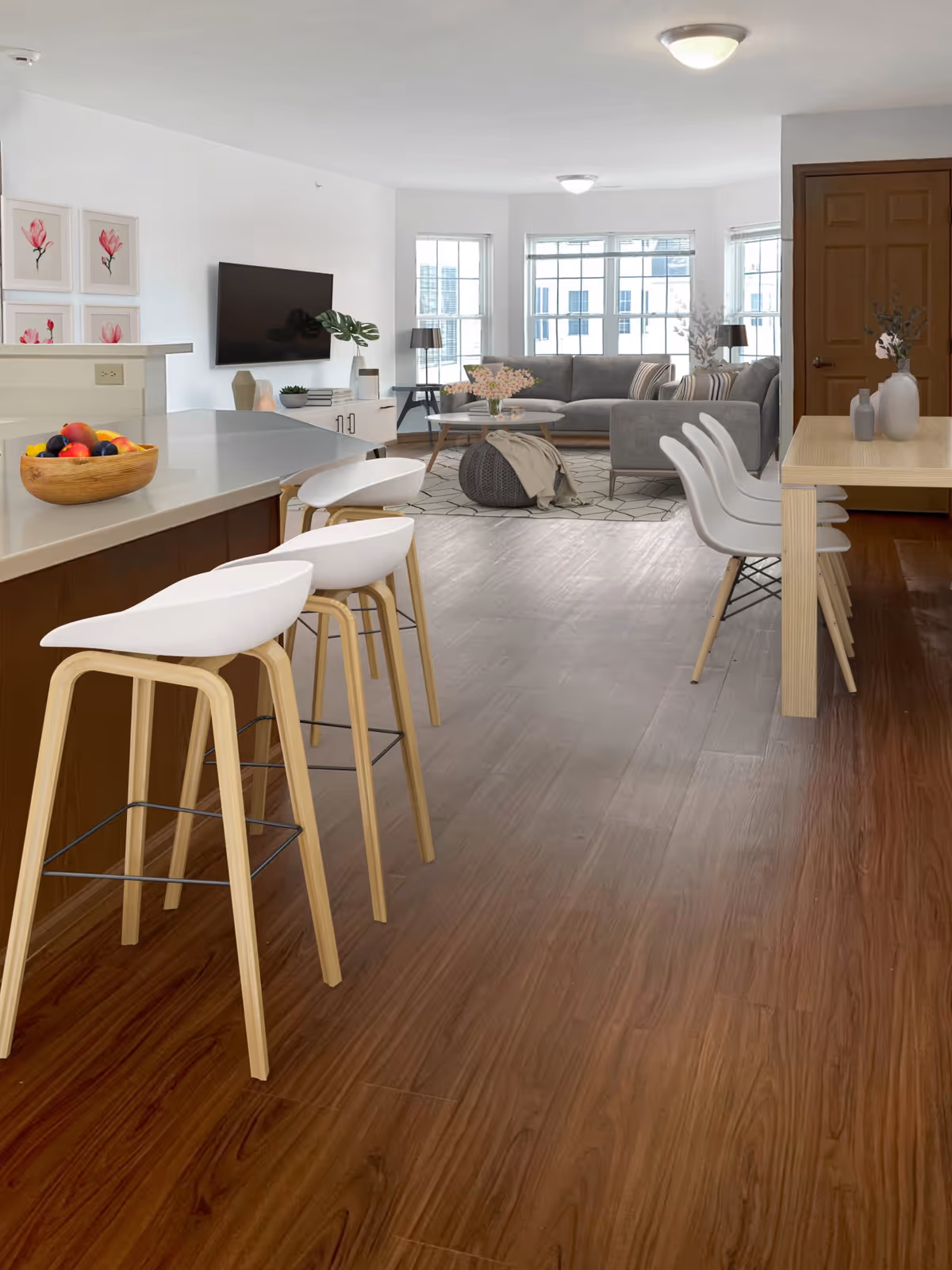 A modern open-concept living space featuring a kitchen island with three white bar stools, a wooden dining table with four white chairs, and a living room area with a gray sectional sofa, a round coffee table, a pouf, and large windows letting in natural light. The walls are white with floral artwork and a mounted flat-screen TV.