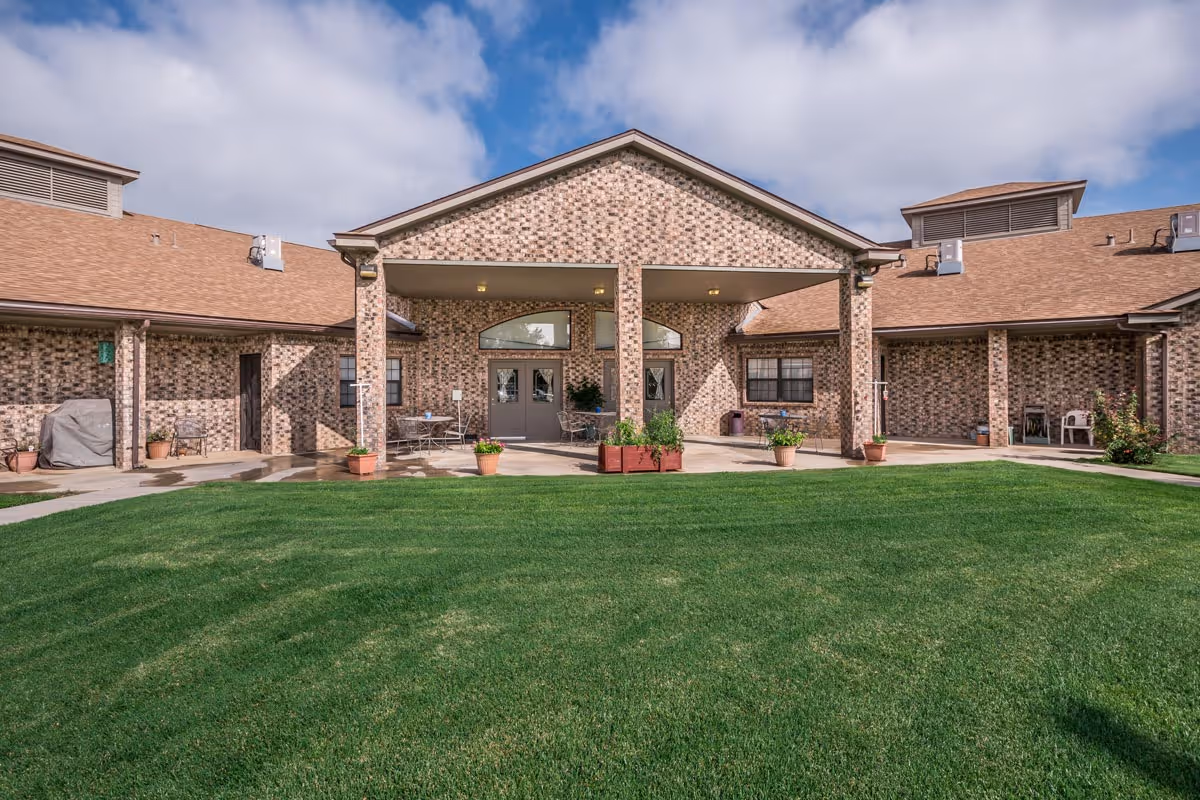 Exterior view of Prairie Acres Nursing Home/Friona Heritage Estates showing a brick building with a covered entrance, several potted plants, and a well-maintained green lawn under a partly cloudy sky.