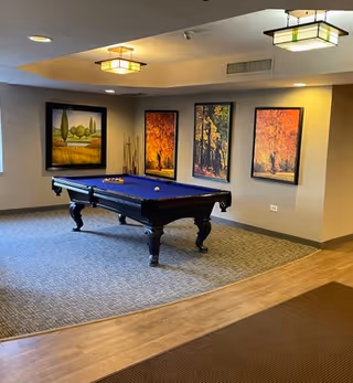 A recreational room with a blue felt pool table centered on a carpeted area. The walls are decorated with four large framed paintings depicting autumn and nature scenes. The ceiling has recessed lighting and two square pendant lights. The floor around the carpeted area is wood, and there is a window on the left side letting in natural light.