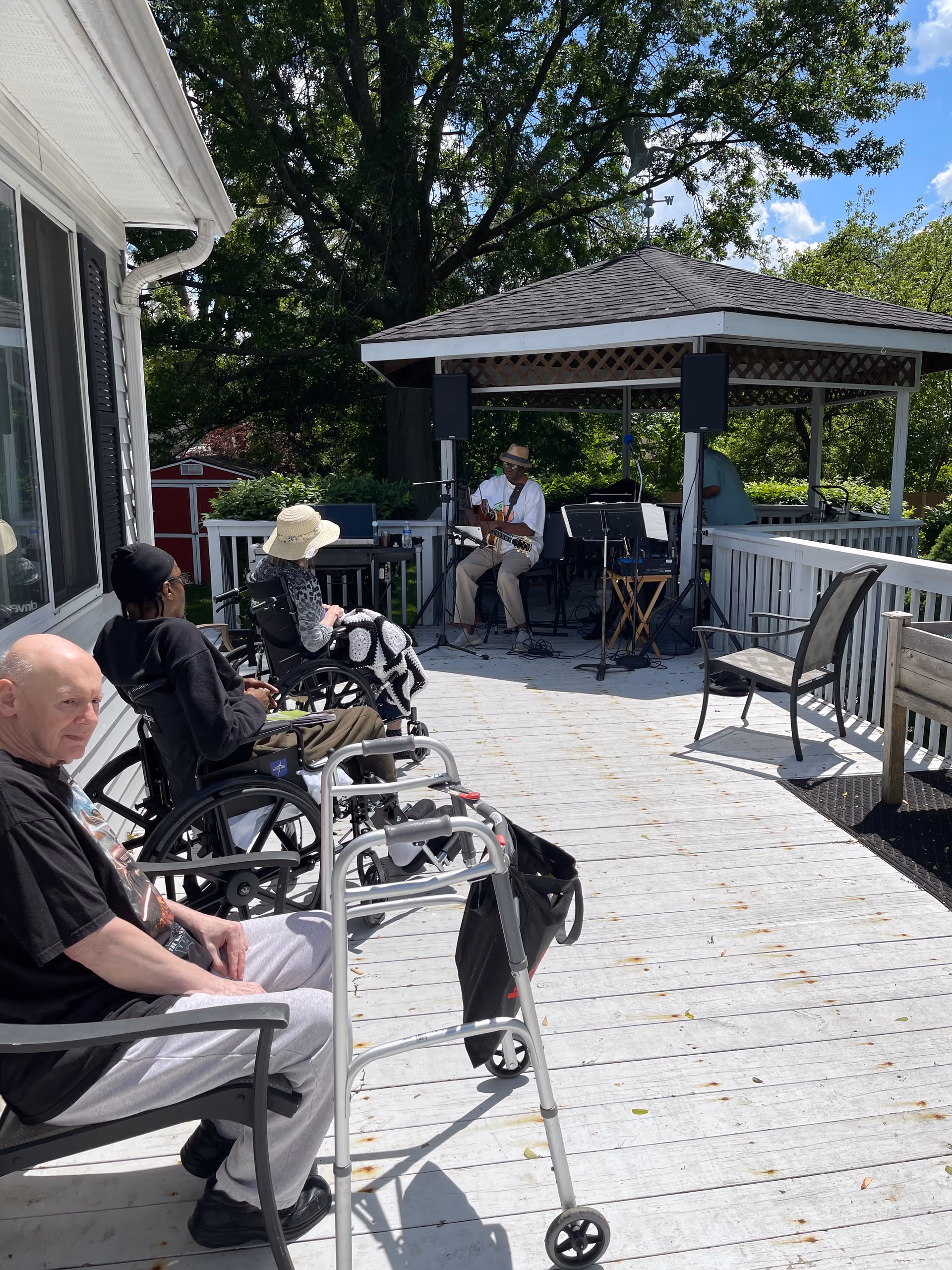 Several elderly individuals sitting on chairs and in wheelchairs on a wooden deck outside a building, listening to a musician playing guitar under a gazebo surrounded by trees and greenery on a sunny day.