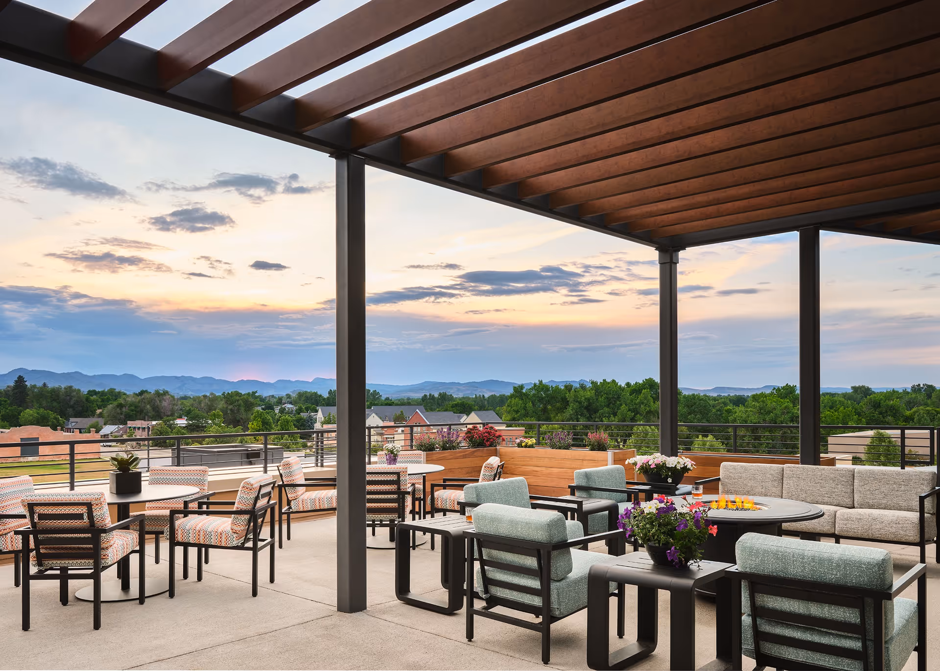 Outdoor patio area with modern seating including cushioned chairs and sofas arranged around tables and a fire pit under a wooden pergola, overlooking a scenic view of trees, rooftops, and distant mountains at sunset.