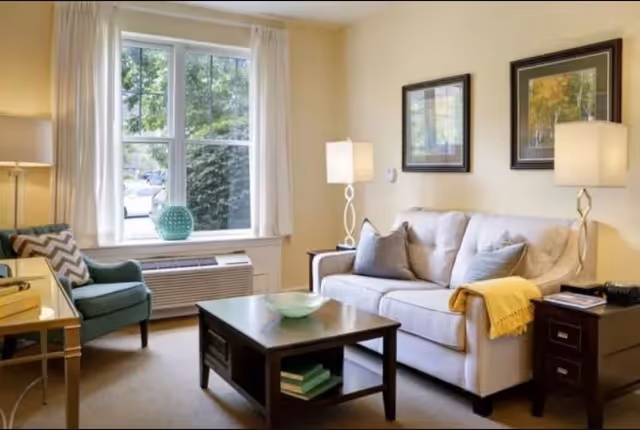 A cozy living room with a beige sofa adorned with gray pillows and a yellow throw blanket. There are two side tables with lamps on either side of the sofa. A dark wooden coffee table with a decorative bowl and books underneath sits in front of the sofa. To the left, there is a blue armchair with a chevron-patterned pillow next to a window with white curtains. The walls are painted light yellow and have two framed pictures hanging above the sofa.