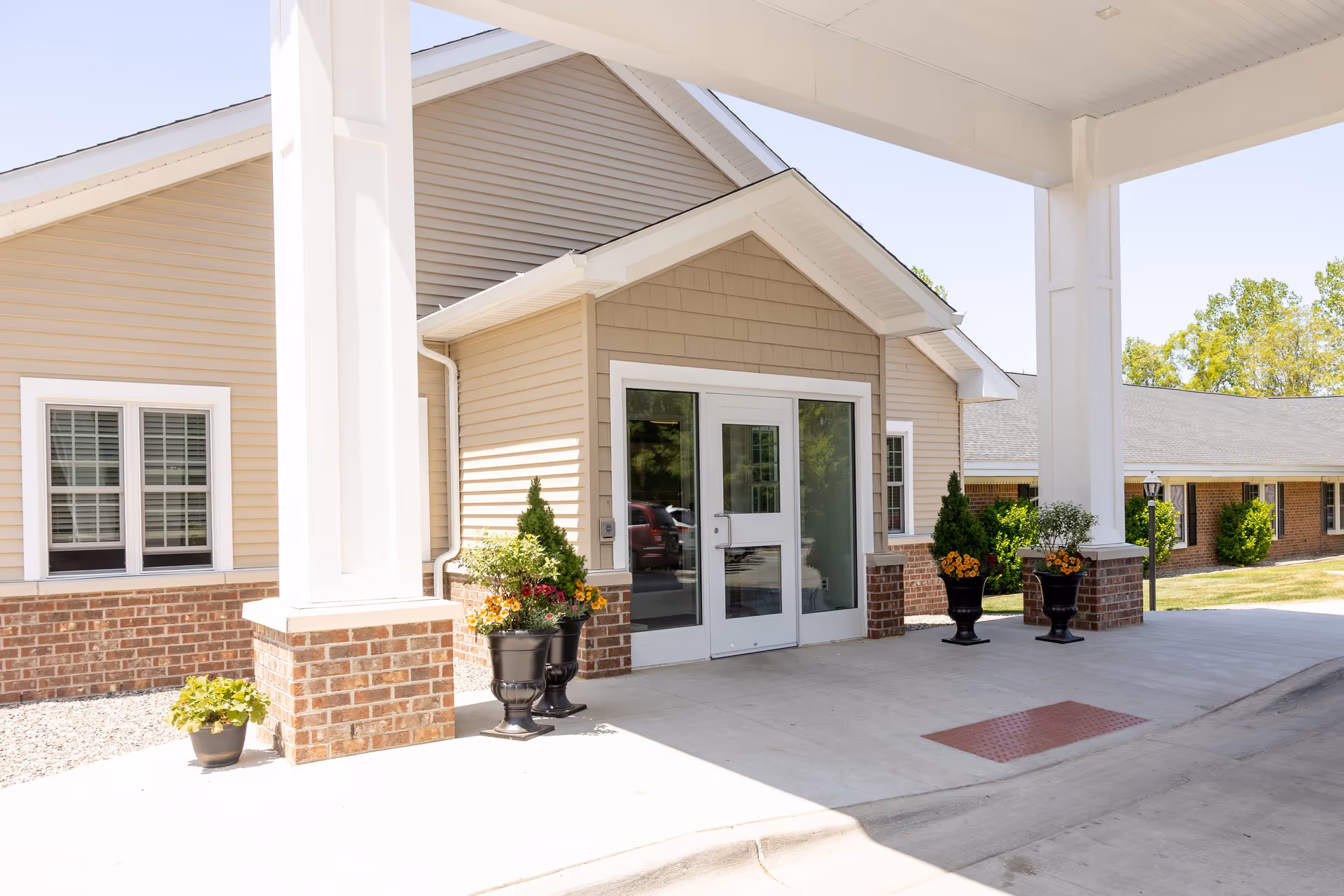 Entrance of a senior living facility with beige siding and brick accents, featuring double glass doors under a covered driveway. There are decorative black planters with flowers and greenery on either side of the entrance, and a small potted plant near a white column. The sky is clear and trees are visible in the background.