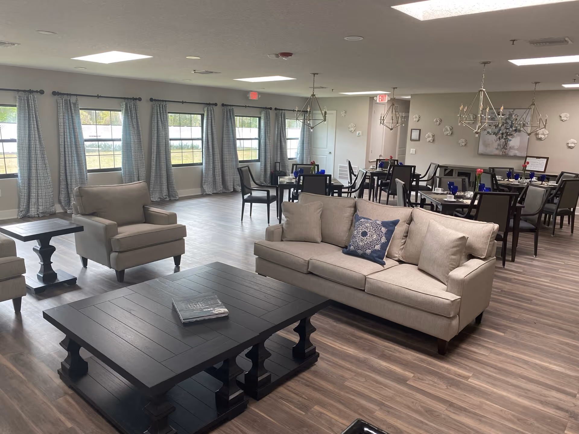 A spacious and well-lit common area featuring a beige sofa with cushions, two matching armchairs, and a large dark wooden coffee table. In the background, there are several dining tables with chairs, blue glassware, and small flower vases. Large windows with light blue curtains allow natural light to fill the room. The walls are decorated with a painting and small white wall sculptures, and modern chandeliers hang from the ceiling.