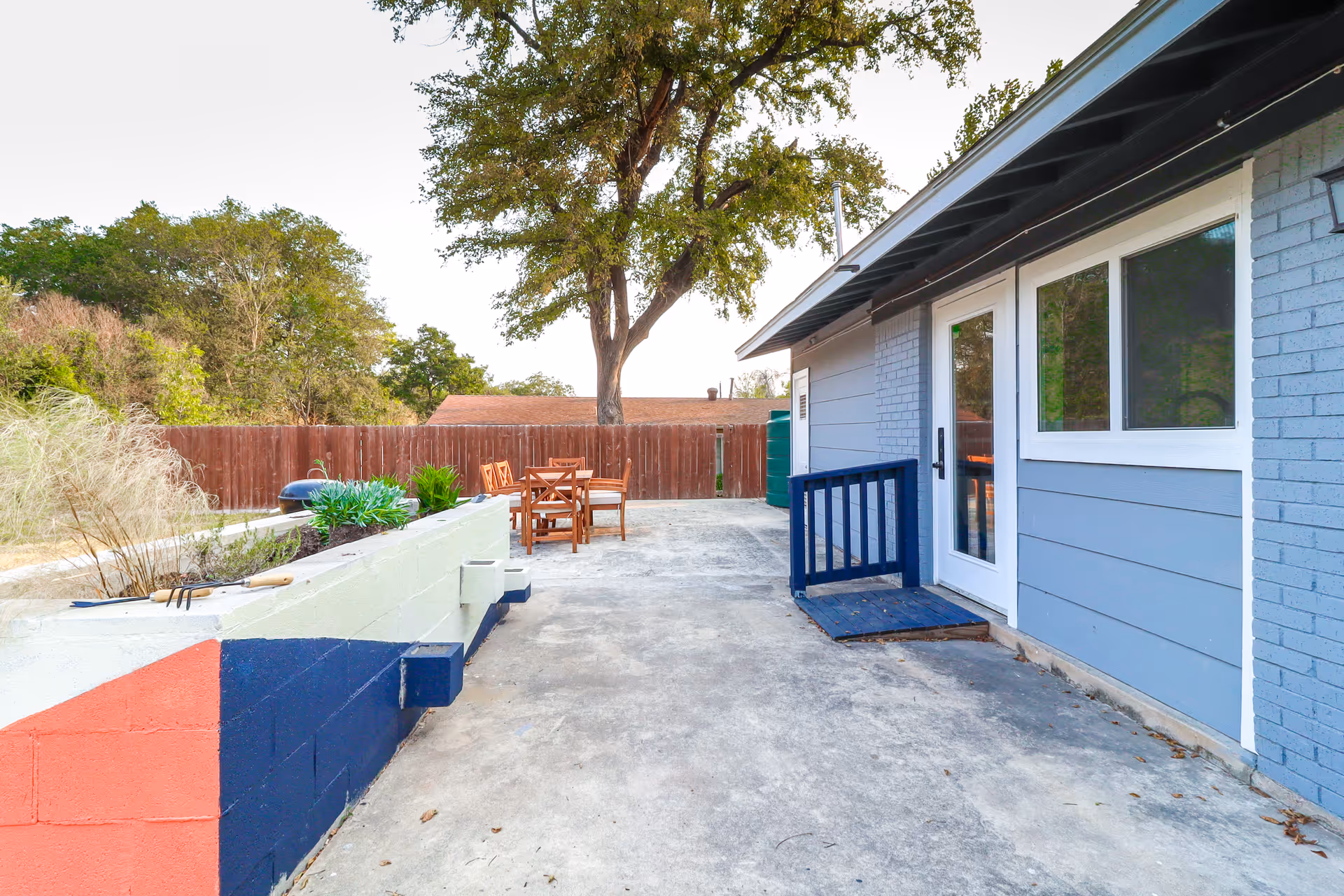 Outdoor patio area at Austin Gardens Senior Living with a concrete floor, a blue and white building wall with a door and window, a small blue ramp, a wooden table with chairs, and a large tree in the background surrounded by a wooden fence.