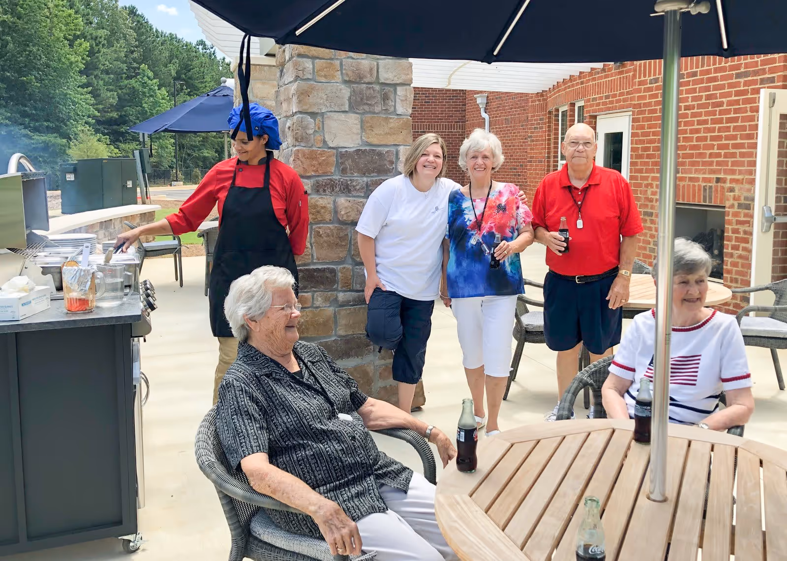 A group of elderly people and a staff member enjoying an outdoor barbecue at Silver Comet Senior Village. The staff member is cooking on a grill while the seniors are sitting and standing around a patio table with drinks, smiling and socializing under large umbrellas.