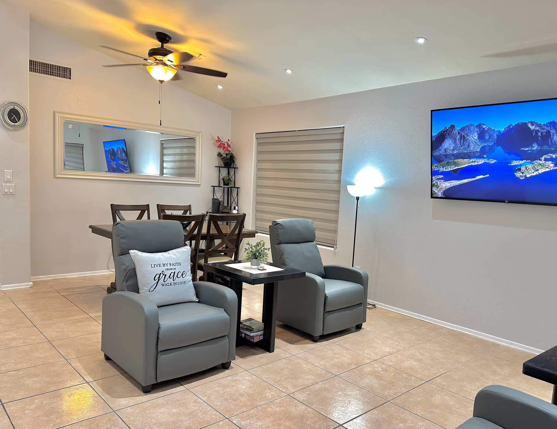A cozy living room area with two gray recliner chairs separated by a small black table. One chair has a decorative pillow with the words 'Live by faith, grow in grace, walk in love.' Behind the chairs is a dining table with four chairs, a large wall mirror, and a small shelving unit with plants and decor. A flat-screen TV is mounted on the wall displaying a scenic mountain and lake view. The room has tiled floors, a ceiling fan with light, a floor lamp, and a window with a beige roller shade.