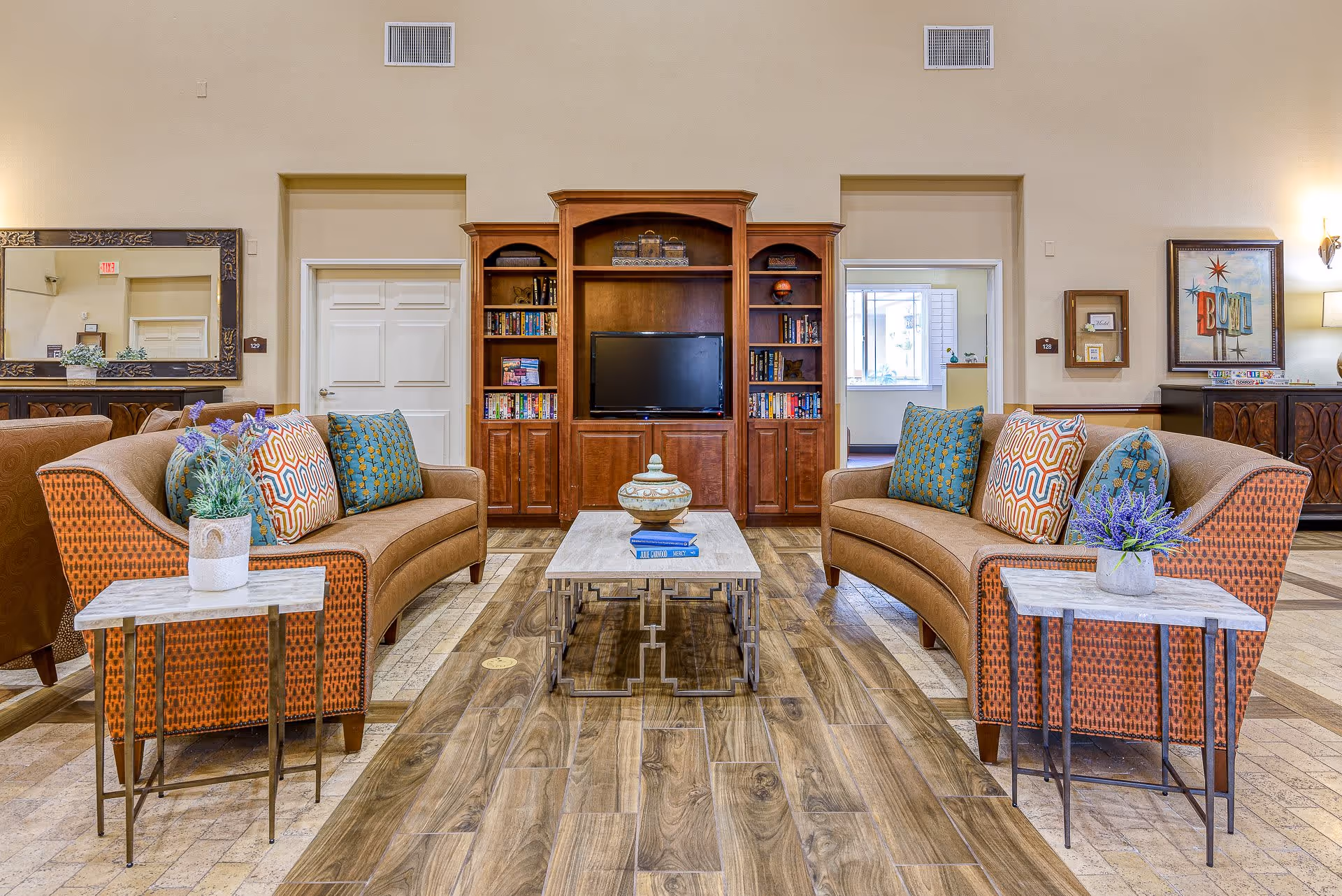 A bright common living room with two curved sofas facing a wooden entertainment center and TV, a central coffee table, and side tables with potted plants.