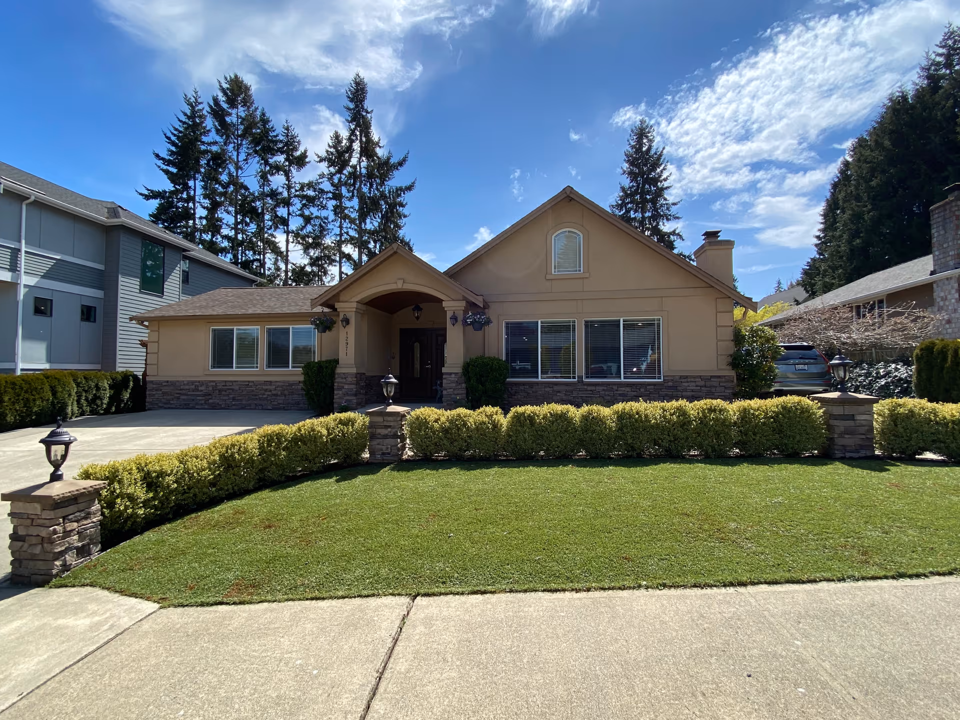 Front exterior view of a single-story residential building with beige walls and stone accents, a well-maintained lawn, trimmed bushes, and a driveway. The sky is partly cloudy with tall evergreen trees in the background.