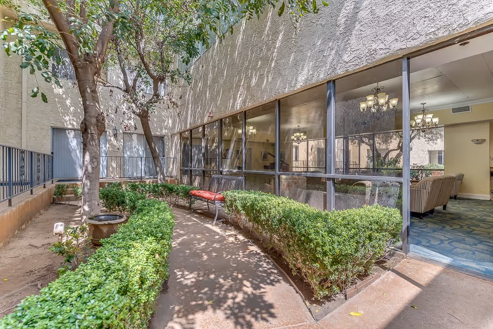 Outdoor courtyard area with a curved pathway bordered by green hedges and a tree. There is a bench with red cushions along the pathway. Large windows show an interior lounge area with chandeliers and seating visible inside the building.