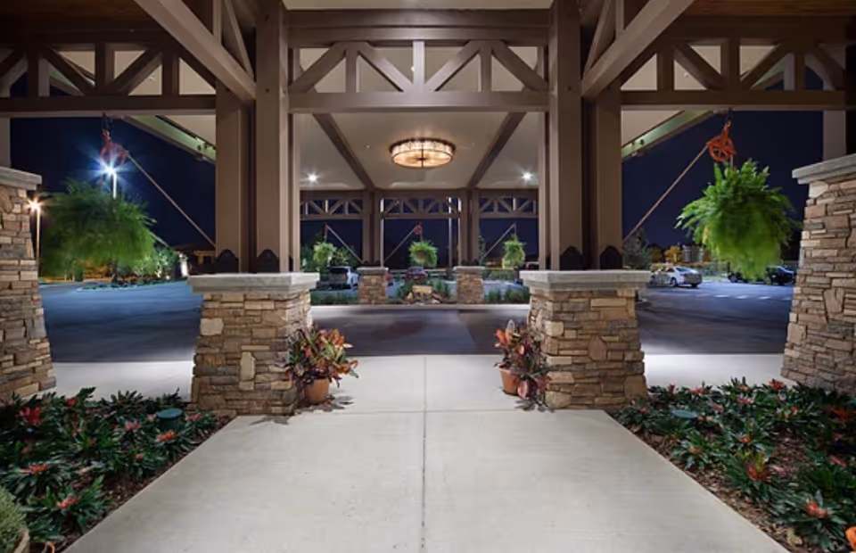 Night view of the entrance area of a facility with stone pillars supporting a wooden roof structure. Hanging green plants are suspended from the ceiling, and there are potted plants and landscaped greenery on either side of the walkway. The parking lot and some parked cars are visible in the background under artificial lighting.