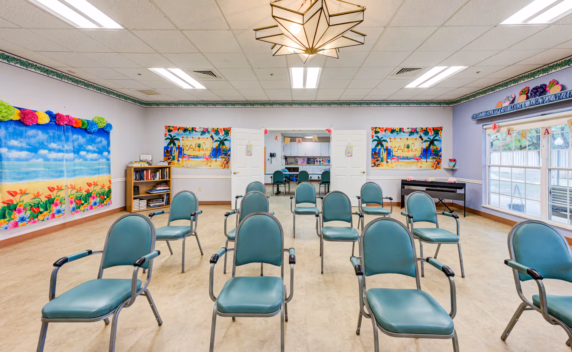 A brightly lit room with several green chairs arranged in rows facing forward. The walls are decorated with colorful tropical-themed posters and garlands. There is a bookshelf with books and games on the left side, and a piano on the right side near large windows letting in natural light. Double doors at the back lead to another room with more chairs and tables.