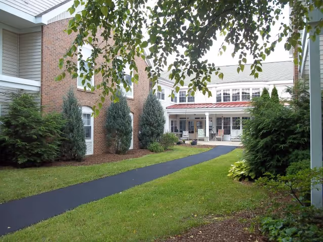 Outdoor view of a senior living facility courtyard with a paved walkway, green grass, shrubs, and trees. The building has brick and white siding with multiple windows and a covered porch area with chairs.