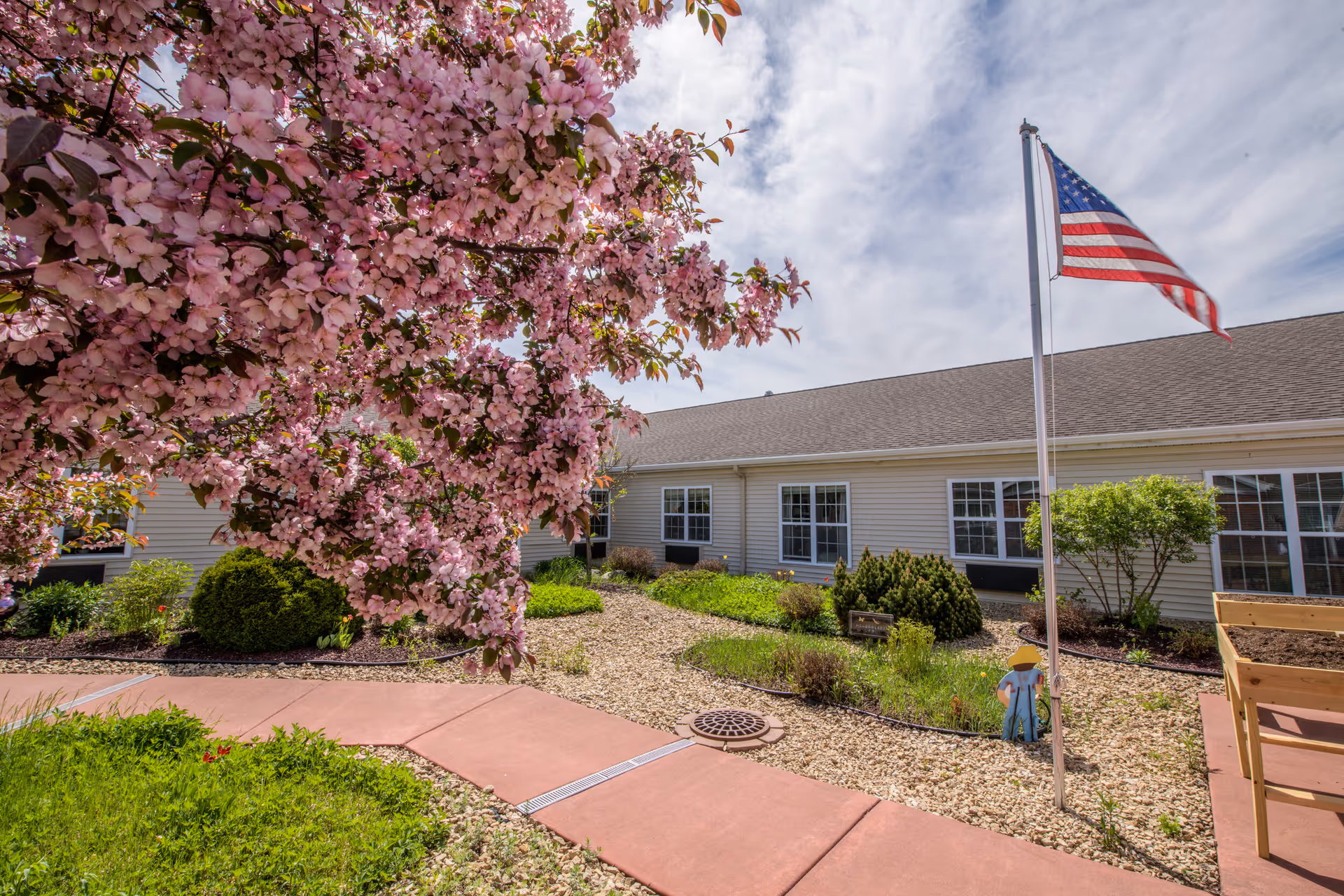 A landscaped outdoor garden area with a blooming tree with pink flowers in the foreground, a paved walkway, green shrubs, and an American flag on a flagpole. The background shows a single-story building with multiple windows under a partly cloudy sky.