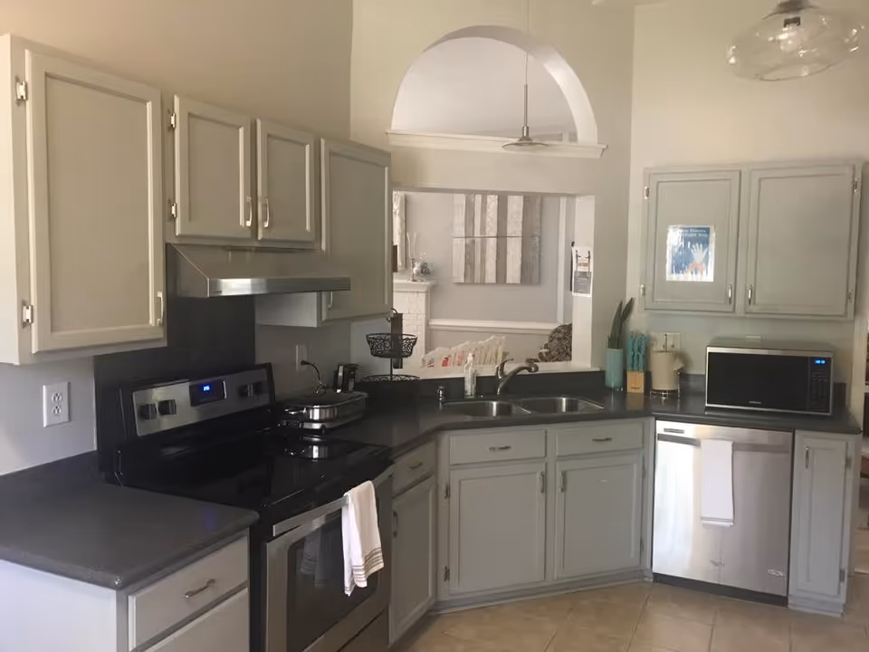 A kitchen area with light gray cabinets, a stainless steel electric stove with a white towel hanging on the handle, a double sink, a microwave, and a dishwasher. The kitchen has dark countertops and beige tiled flooring. There is an open archway above the sink that looks into a living room area with a white fireplace and wall art.