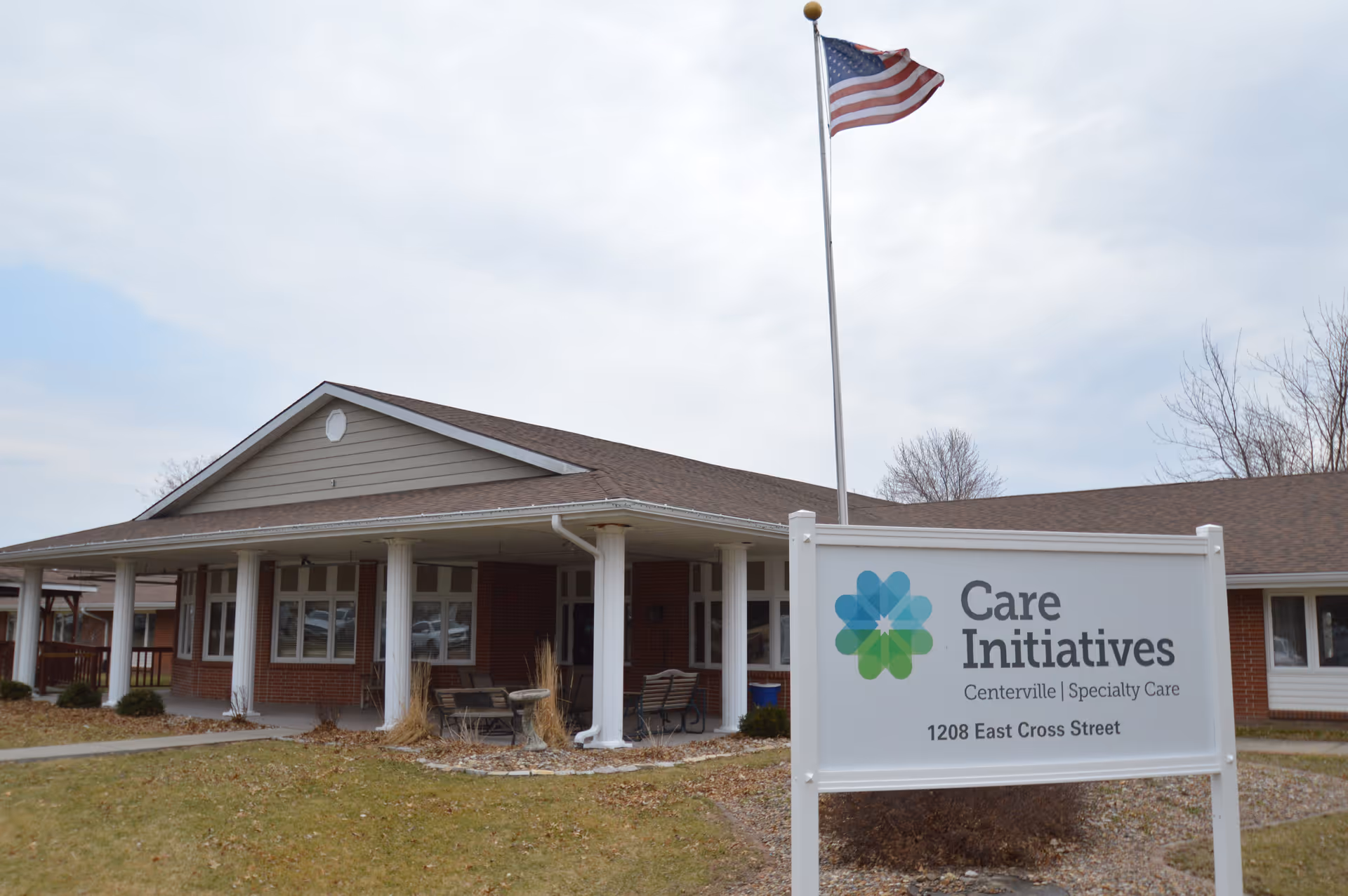Exterior view of Care Initiatives Centerville Specialty Care building with a covered porch, several windows, and an American flag on a flagpole. A sign in front displays the facility name and address: 1208 East Cross Street.