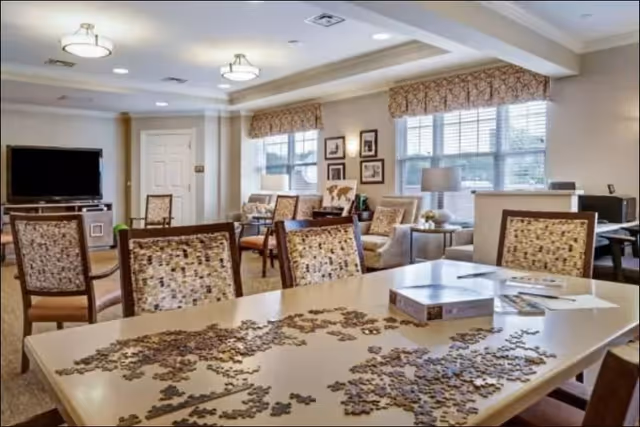 A well-lit common area with a table in the foreground covered with puzzle pieces and a puzzle box. Surrounding the table are several cushioned chairs with patterned upholstery. In the background, there are more chairs, a television on a stand, windows with valances, framed pictures on the wall, and a lamp on a side table.