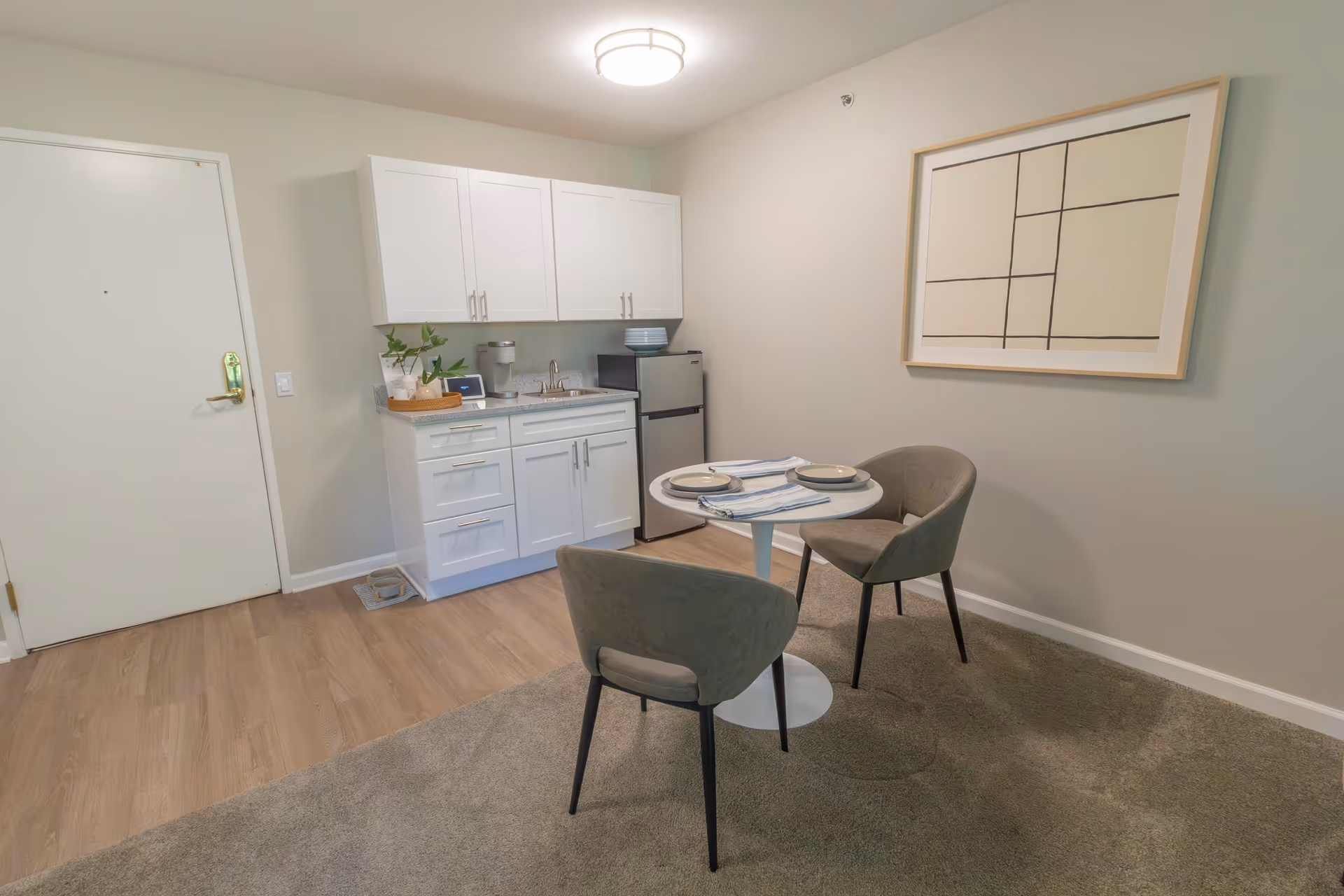 A small dining area with a round white table set for two with plates and napkins, two gray upholstered chairs, a kitchenette with white cabinets, a small sink, a mini fridge, and a decorative plant on the counter. The walls are light-colored with a framed abstract artwork, and the floor is a combination of wood and carpet.
