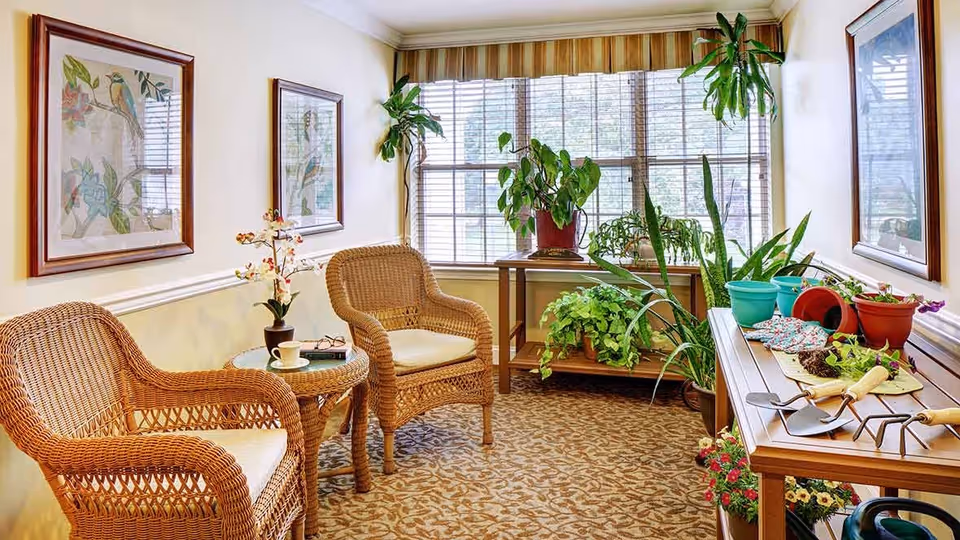 A cozy indoor sitting area with two wicker chairs and a small round table between them holding a cup and a book. The room has a large window with blinds, several potted plants on a wooden table and a plant stand, gardening tools and pots on a wooden bench, and framed botanical artwork on the walls.