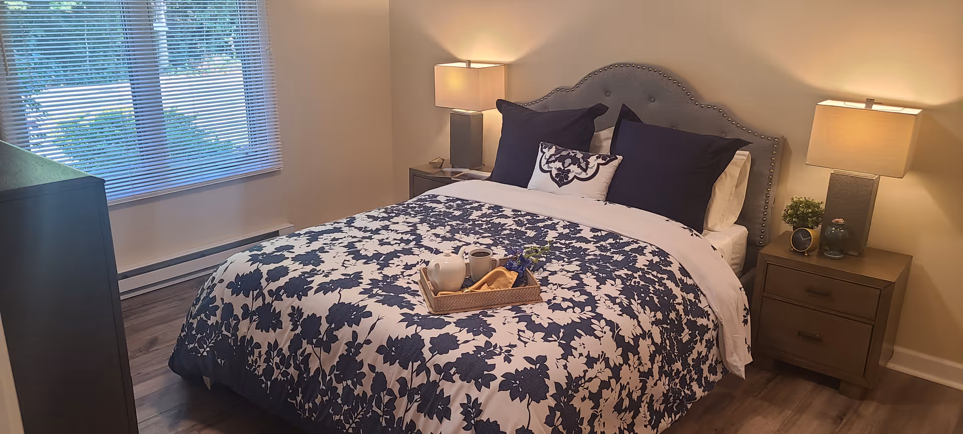 A neatly made bed with a blue and white floral patterned comforter and matching pillows in a bedroom. There are two bedside tables with lamps on each side of the bed, a small plant, and decorative items. A window with blinds is on the left side, and the floor is wooden.