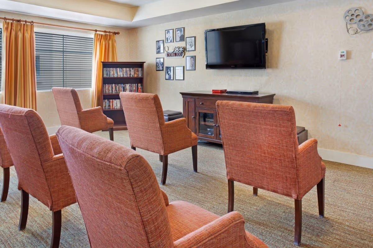 A small media lounge with rows of upholstered chairs facing a wall-mounted TV and media cabinet, a DVD shelf, and a curtained window.
