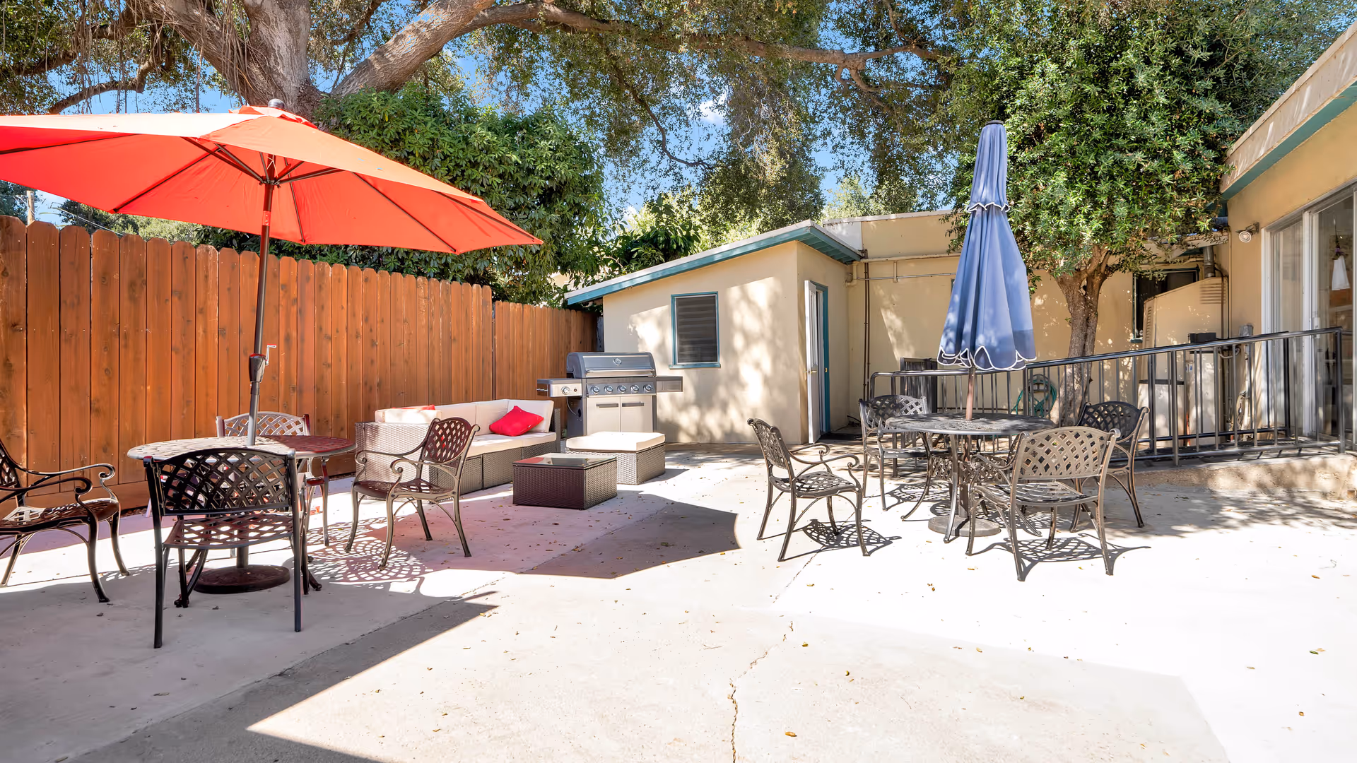 Outdoor patio area with metal tables and chairs, two umbrellas (one red and one blue), a cushioned seating area with a red pillow, a barbecue grill, and surrounding trees and wooden fence under a clear blue sky.