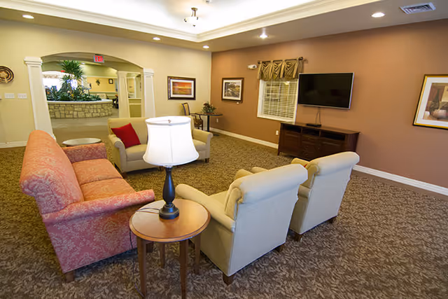 A cozy living room area in a senior living facility with a red patterned sofa, two beige armchairs, a small round wooden table with a lamp, a wall-mounted flat screen TV, framed artwork on the walls, and a window with blinds and a valance. The room has beige walls, carpeted floor, and an open doorway leading to another area with a stone counter and plants.