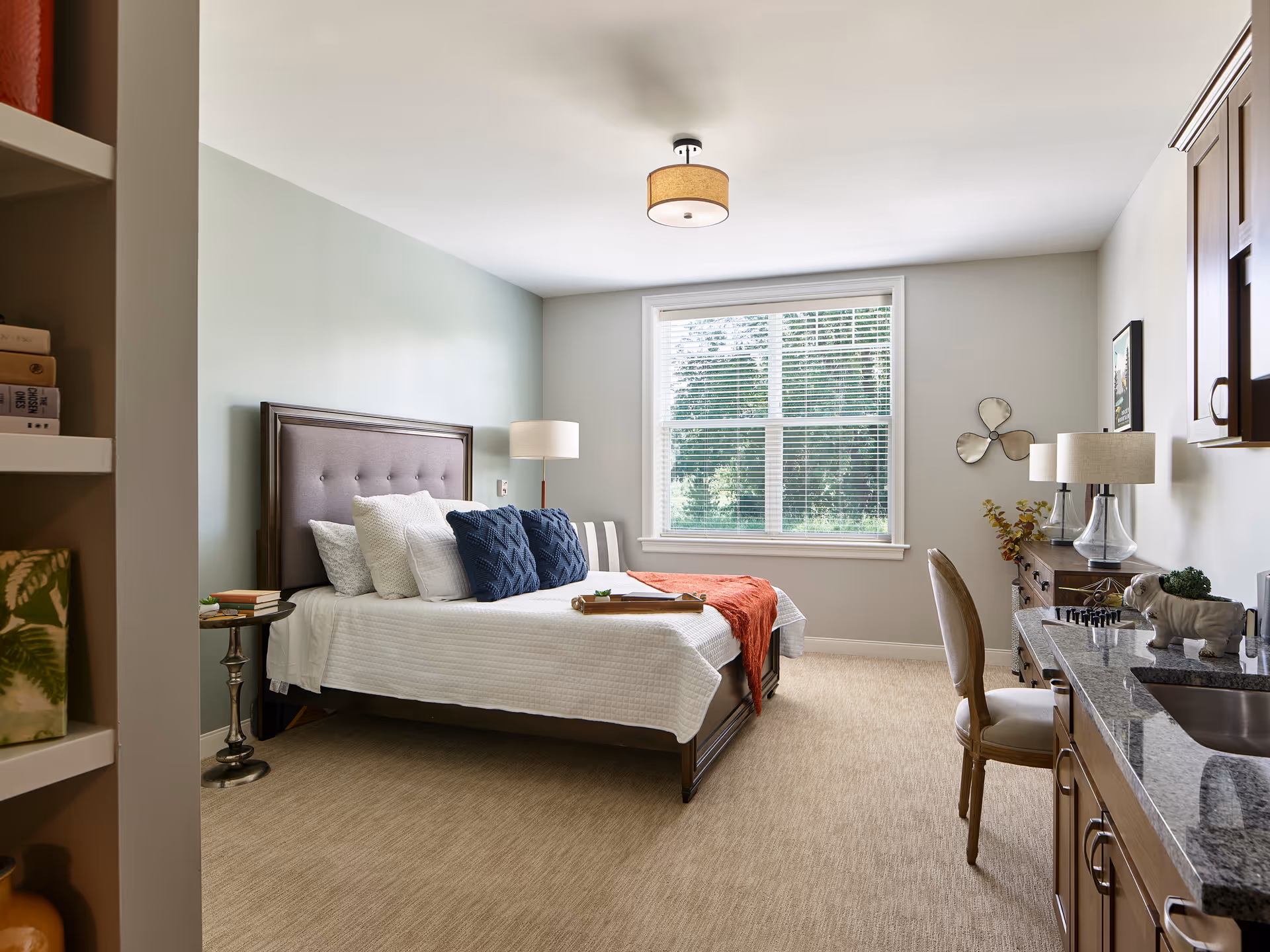 A bright and cozy bedroom with a large window letting in natural light. The room features a bed with white bedding and blue accent pillows, a wooden headboard, a side table with books, a floor lamp, a wooden dresser with a table lamp and decorative items, a chair, and a kitchenette area with a granite countertop and sink. The walls are painted in soft neutral tones and the floor is carpeted.