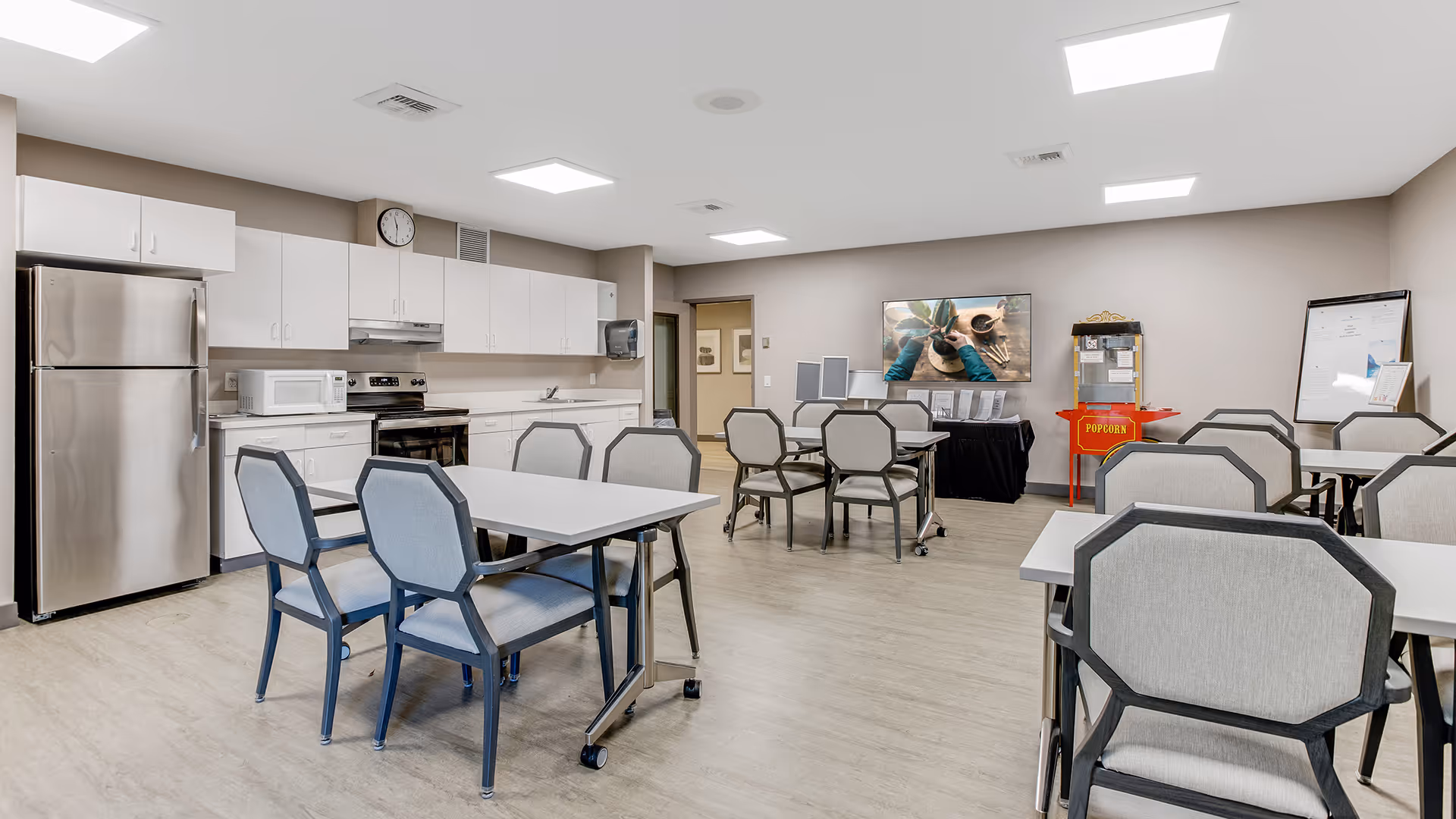 A bright and clean communal dining area with several tables and chairs. The room features a kitchen area with white cabinets, a stainless steel refrigerator, microwave, and stove. On the right side, there is a popcorn machine and a bulletin board. The walls are painted light beige, and the floor has a light wood finish.