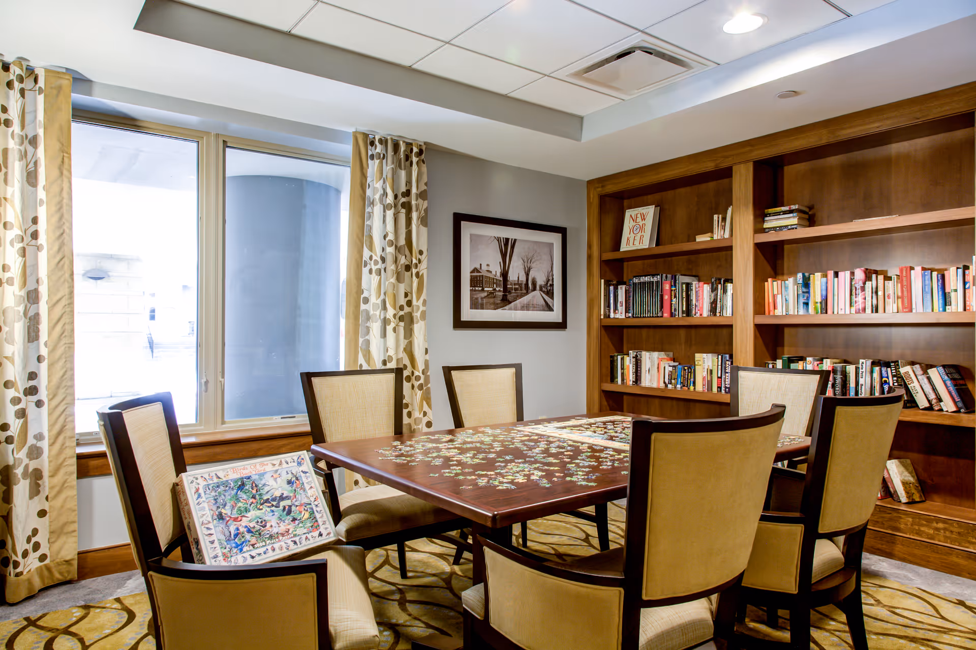 A bright communal activity room with a table and chairs mid-puzzle, bookshelves, and a large window with patterned curtains.