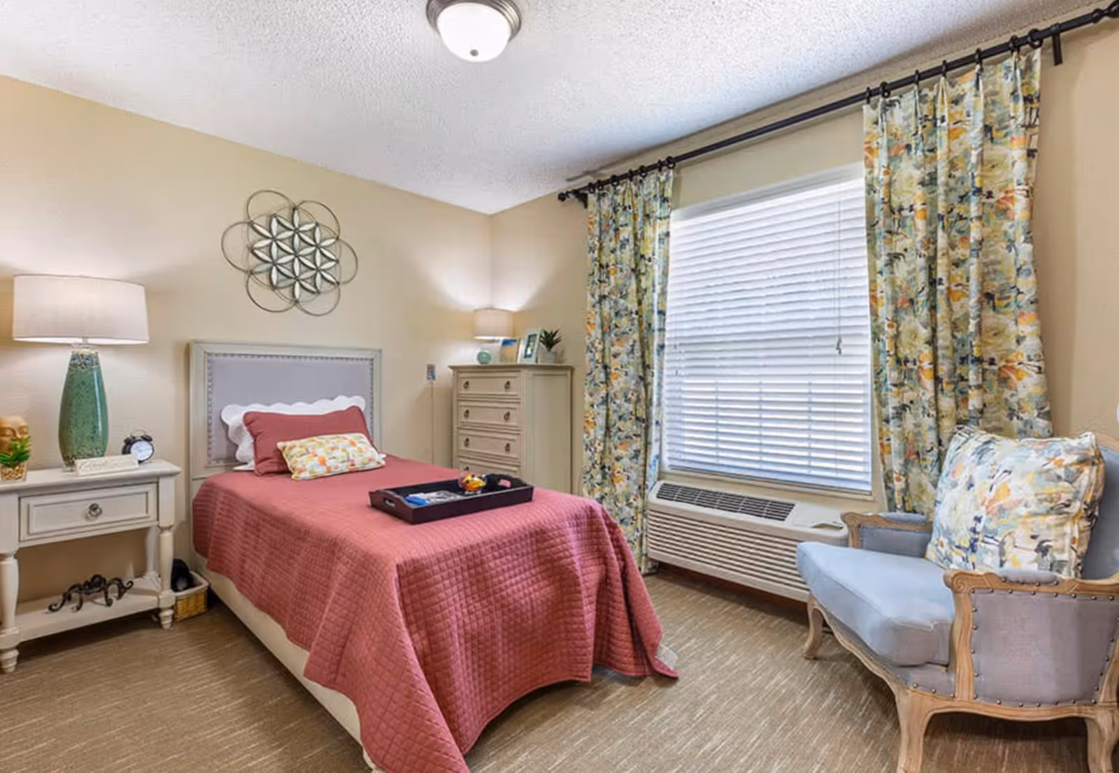 Sunlit furnished bedroom with a single bed covered in a pink quilt, nightstand and dresser, floral curtains, and an upholstered armchair.