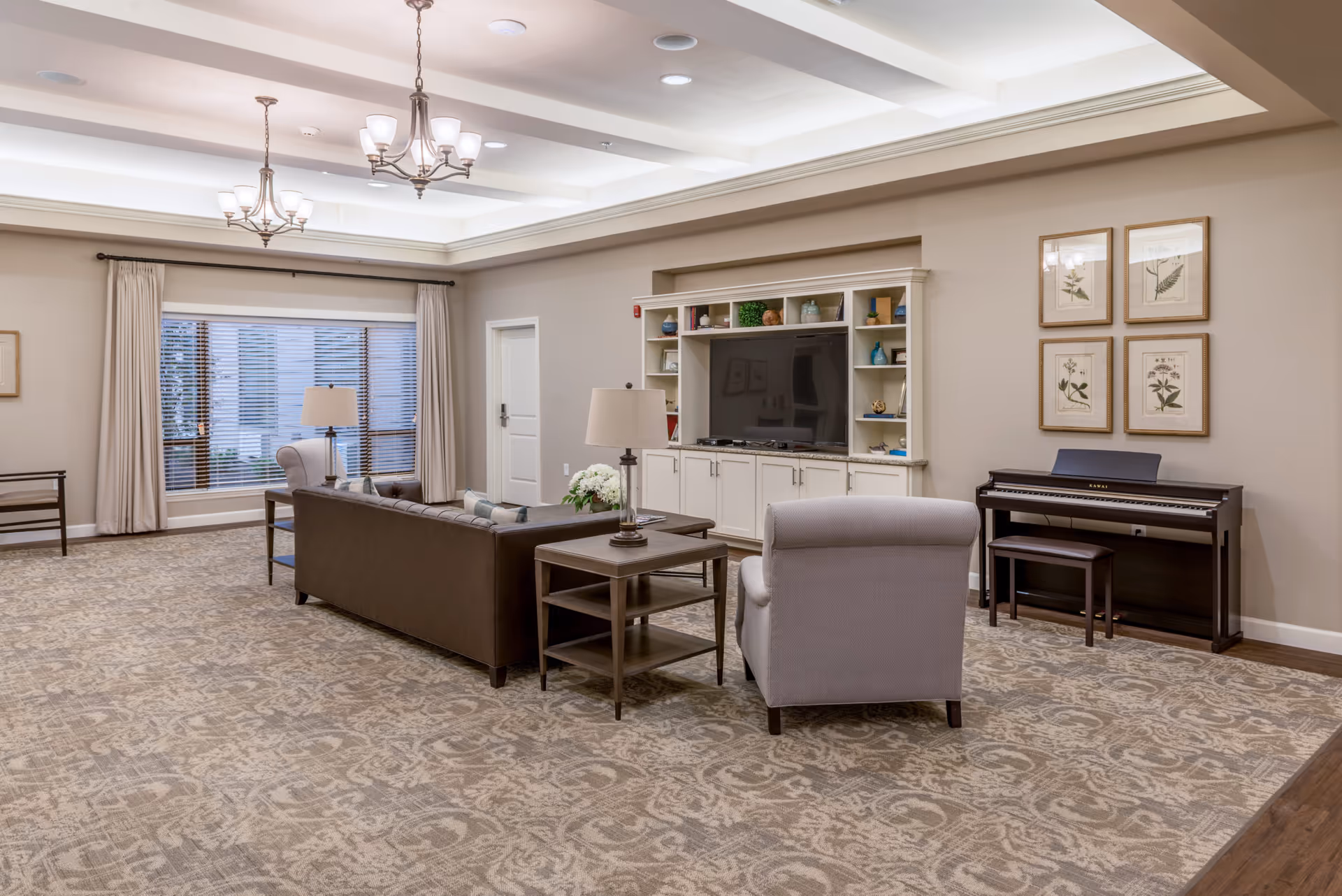 A spacious senior living room with a patterned carpet, a brown leather sofa, a gray armchair, two side tables with lamps, a large flat-screen TV on a white built-in cabinet with shelves, a digital piano against the wall, and framed botanical prints above the piano. The room has large windows with blinds and curtains, and two ceiling chandeliers.
