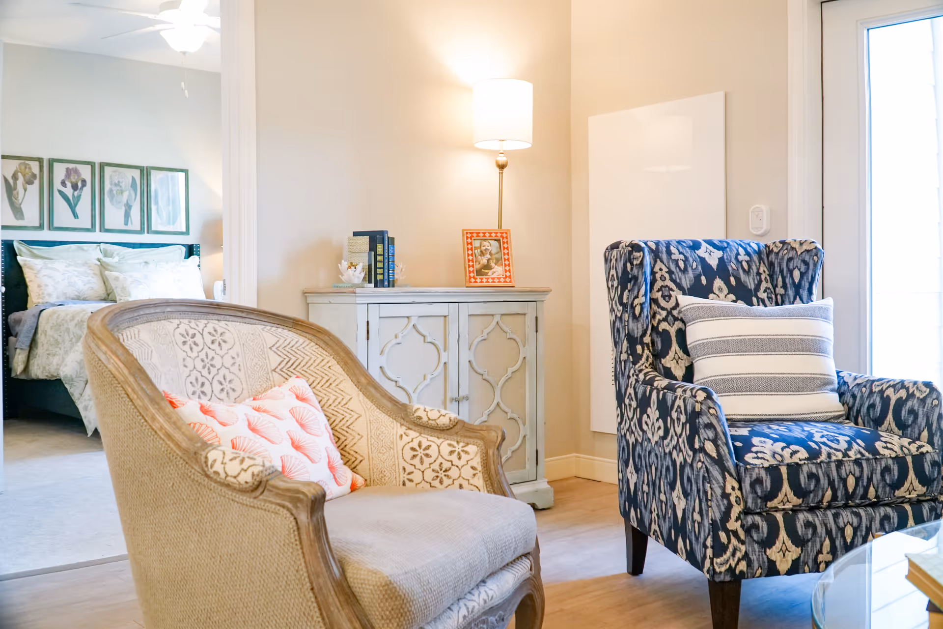 A cozy interior living space featuring two armchairs, one beige with patterned upholstery and a coral-patterned pillow, and the other blue with a white and gray striped pillow. Behind the chairs is a small white cabinet with decorative details, topped with books, a framed photo, and a lamp. In the background, a bedroom with a bed, pillows, and floral artwork on the wall is visible through an open doorway.