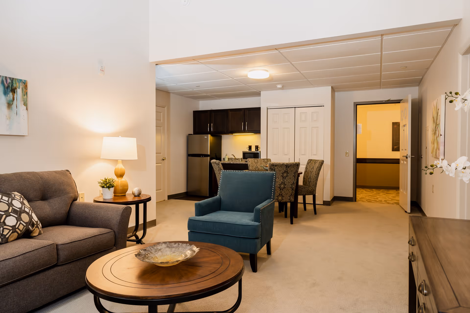 Open living room and dining area with a sofa, blue armchair, round coffee table, and a kitchenette in the background.