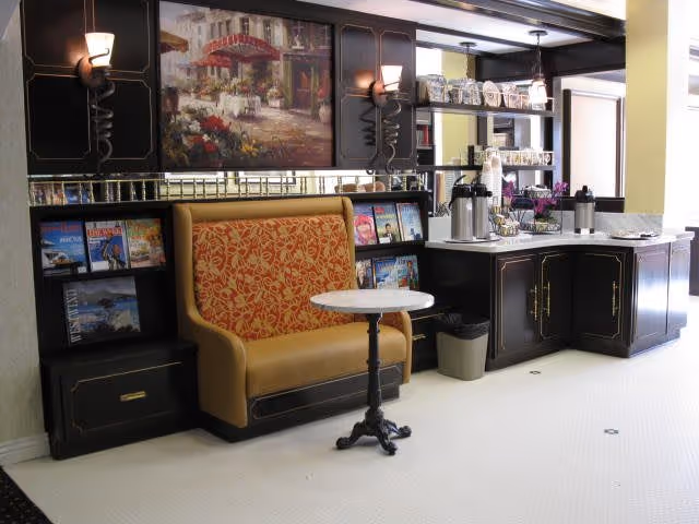 A small seating nook with an upholstered bench, round table, magazine racks, and a self-serve coffee station under framed wall art.
