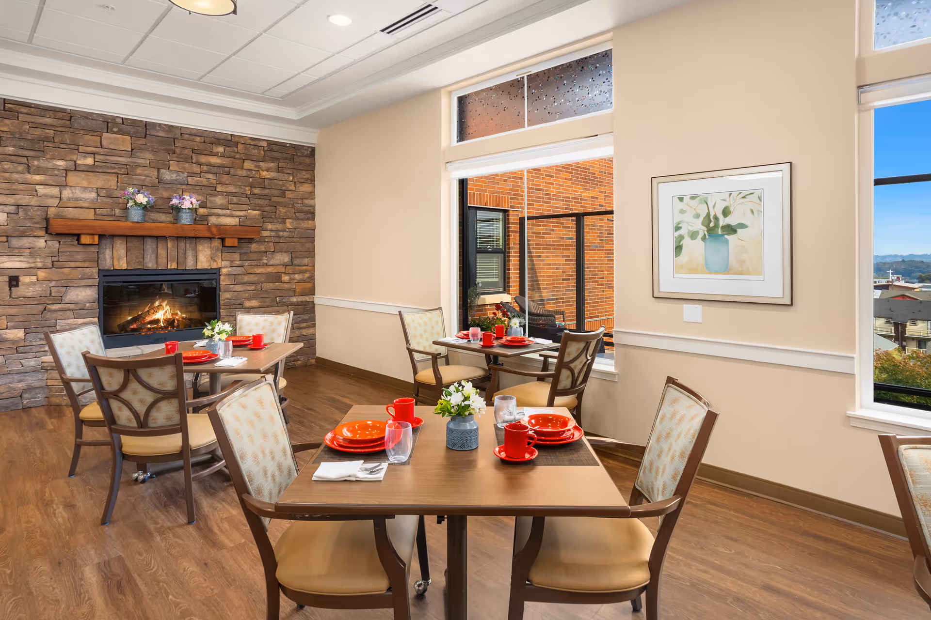 Dining room with multiple set tables and chairs, a lit stone fireplace, and large windows.