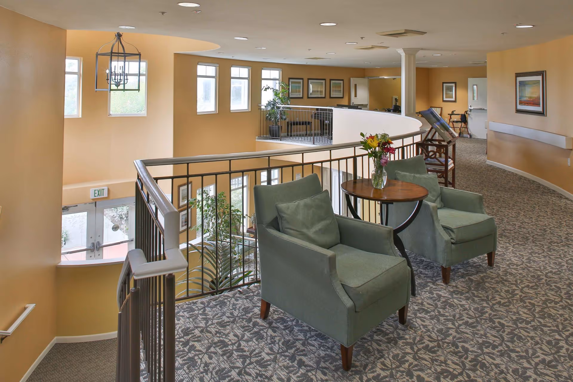 Interior view of a senior living facility hallway with a seating area featuring two green armchairs and a small round wooden table with a vase of flowers. The hallway has patterned carpet, beige walls, framed artwork, and a railing overlooking the lower floor with large windows letting in natural light.