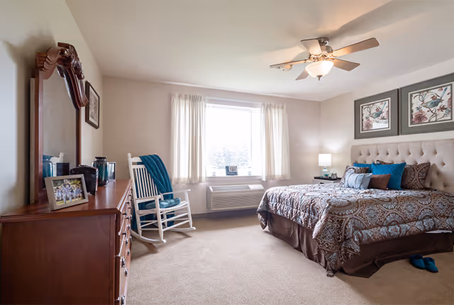 Sunlit furnished bedroom with a tufted bed and patterned bedding, a dresser with mirror, a white rocking chair, and a ceiling fan.